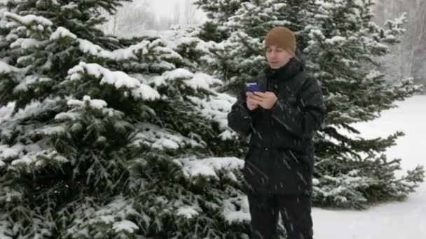 Ung man att skriva ett SMS på telefonen i vinter snöiga skogen. Stora snöfall. Han ler och talar det skriftliga. — Stockvideo