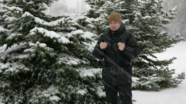 Um jovem no inverno está frio na floresta perto do abeto. Grande queda de neve. Ele está respirando nas mãos, esfrega e usa luvas com um capuz. Paisagem nevada. Ele olha para a câmara. — Vídeo de Stock