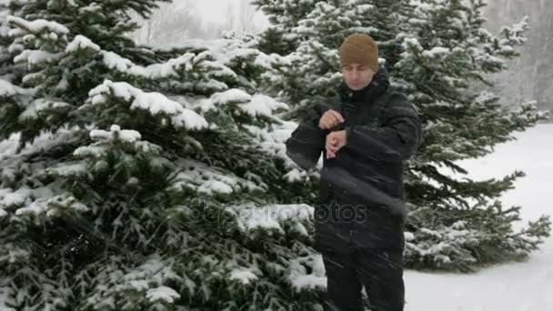 男は、雪に覆われた冬の森でスマートな時計にメッセージをチェックします。大きな降雪。彼は時計に音声応答を決定します。暗い暖かいジャケットに身を包んだ男. — ストック動画