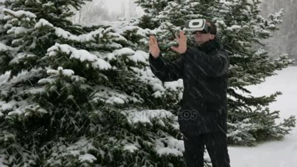 Muž na sobě soupravu pro virtuální realitu v zimě v zasněženém lese. Velké sněžení. Snaží se dotknout virtuální objekty. Muž oblečený do teplé bundě a mává rukama. — Stock video