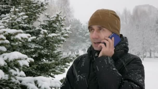 A young man in winter forest talking on the phone. Big snowfall. He admires the sides of snow and trees. A man in a dark jacket and a warm hat. — Stock Video
