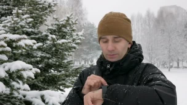 Un uomo controlla i messaggi sull'orologio intelligente nella foresta innevata invernale. Grosse nevicate. Lui detta una risposta vocale all'orologio. Un uomo vestito con una giacca scura e calda . — Video Stock