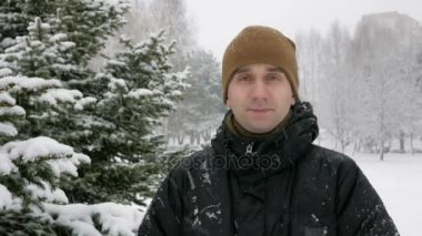 Kamera ve kış ormanda gülümseyen genç adam görünüyor. Büyük kar yağışı. Bir adam bir koyu renk ceketli ve sıcak bir şapka.