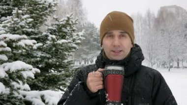 Bir kış ormandaki bir termos sıcak bir içecek içme bir genç adam. Büyük kar yağışı. Adam kameraya doğru görünüyor, koyu renk ceketli ve sıcak bir şapka. doğa gevşeme kavram