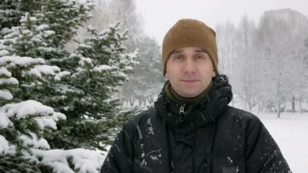 Молодой человек смотрит в камеру и улыбается в зимнем лесу. Большой снегопад. Человек в тёмной куртке и тёплой шляпе . — стоковое видео