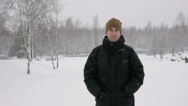 El joven mira a la cámara y sonríe en el bosque invernal. Grandes nevadas. Un hombre con una chaqueta oscura y un sombrero caliente . — Vídeos de Stock