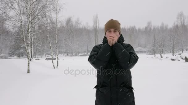 Kışın genç bir adam ormanda soğuktur. Büyük kar yağışı. Ellerinin üstünde nefes hes losyonları ve bir kukuleta ile eldiven giyer. Karlı manzara. Kameraya bakıyor — Stok video