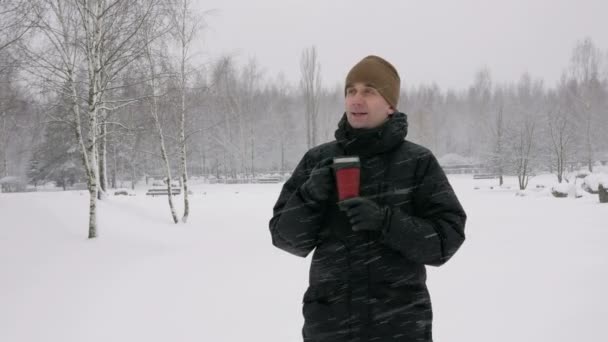 Молодий чоловік п'є гарячий напій з термоса в зимовому лісі. Великий снігопад. Чоловік дивиться в камеру, одягається в темну куртку і теплий капелюх. концепція релаксації на природі — стокове відео