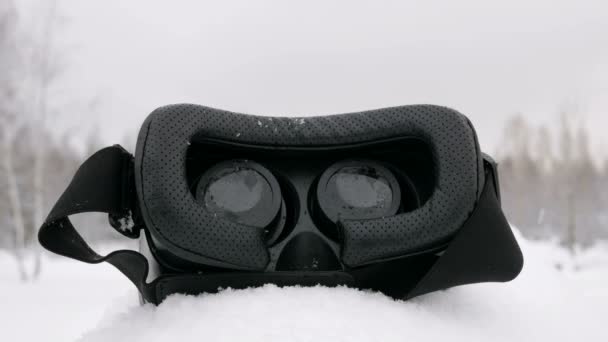 Гарнитура Virtual Reality лежит на снегу в лесу. Снег идёт. Объектив к камере — стоковое видео