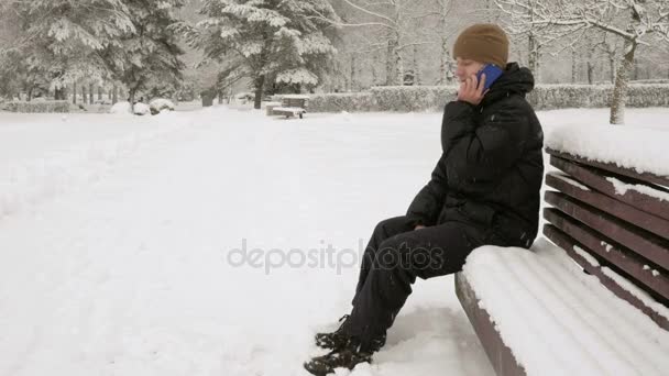 Un joven en el parque de invierno hablando por teléfono. Admira los lados de la nieve y los árboles. Un hombre con una chaqueta oscura y un sombrero caliente . — Vídeo de stock