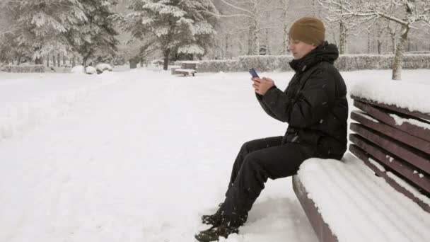 Молодой человек набирает смс по телефону в зимнем снежном парке. Он улыбается и говорит написанное . — стоковое видео