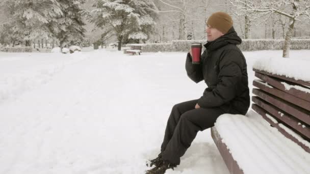 Молодий чоловік п'є гарячий напій з термоса в зимовому парку. Великий снігопад. Чоловік у темній куртці і теплий капелюх. концепція релаксації на природі — стокове відео