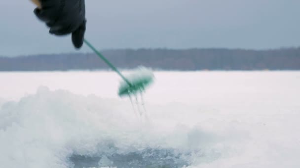 El hombre se dedica a la pesca de invierno en un lago congelado. Limpia el agujero en la corteza de hielo . — Vídeo de stock