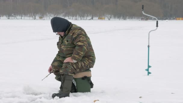 El hombre se dedica a la pesca de invierno. Cuesta alrededor de cañas de pescar esperando peces. Vestido con ropa protectora — Vídeos de Stock