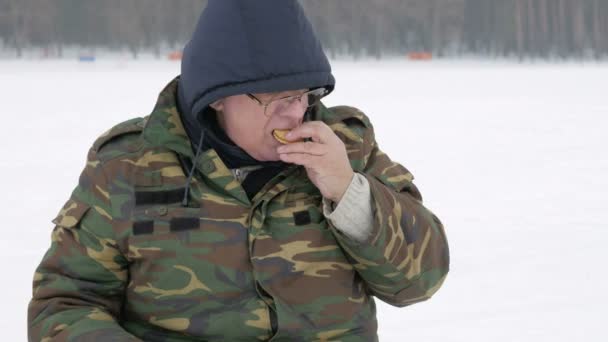 Um homem pescando no lago no inverno e bebendo chá de uma garrafa térmica. Ele come biscoitos com uma bebida quente — Vídeo de Stock