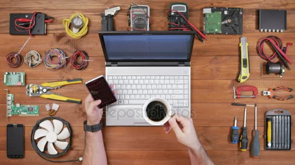 Ingegnere informatico uomo che lavora ad una scrivania con un telefono, un computer portatile e una tazza di caffè. Tavolo in legno vista dall'alto. Controlla il messaggio e guarda la foto — Video Stock