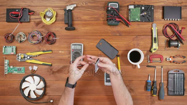 Man reparera en mobiltelefon. Kontrollerar delar inuti enheten. Trä table top-översikt. — Stockvideo