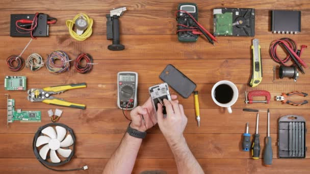 Um homem a reparar um telemóvel. Verifica partes dentro do dispositivo. Tabela de madeira vista superior . — Vídeo de Stock