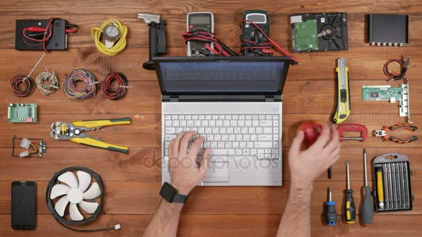 Инженер-мужчина печатает за ноутбуком и ест яблоко. Деревянный стол для ремонта электроники вид сверху — стоковое видео
