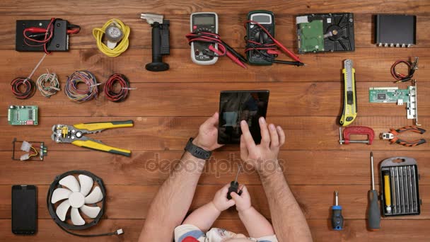 Ingeniero masculino con su hijo mirando foto en la placa de la tableta. Chico sosteniendo un destornillador en su mano. Mesa de madera vista superior — Vídeo de stock
