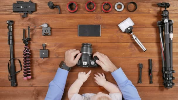 O fotógrafo pai com um jovem filho verificou a câmera antes de filmar. Mesa de madeira vista superior. A criança a tocar num botão. Equipamento para fotografar na mesa . — Vídeo de Stock