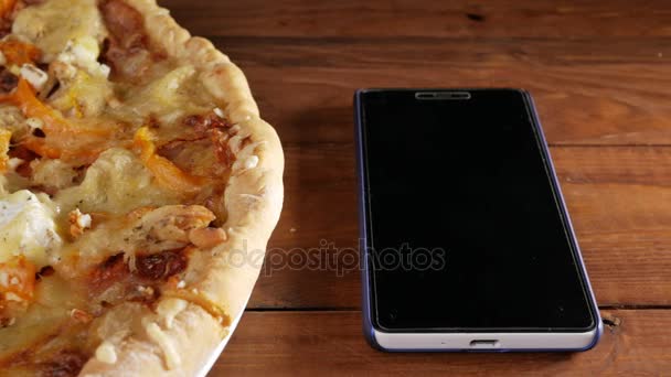 El teléfono móvil descansa sobre una mesa de madera junto a una pizza casera con diferentes quesos y tomate. Cámara móvil — Vídeos de Stock