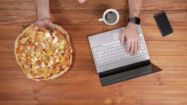 Een man achter een laptop werkt en eet pizza. Vlakbij zijn er is een koffiemok. Houten tafelblad weergave — Stockvideo