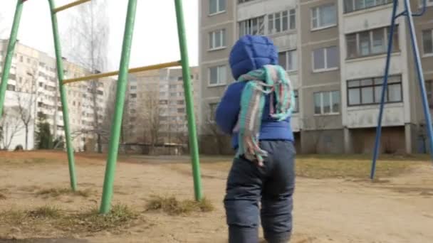 Un chico atractivo ha estado caminando con su madre durante 2 años en el patio de recreo a principios de la primavera. Vestido con una chaqueta azul y sombrero . — Vídeos de Stock