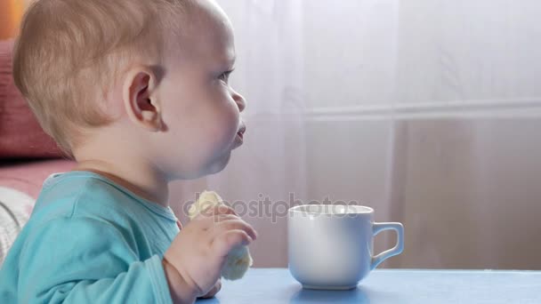 Um menino atraente de 2 anos come uma banana. Senta-se à mesa em casa. Cuidadosamente assistindo desenhos animados na TV . — Vídeo de Stock