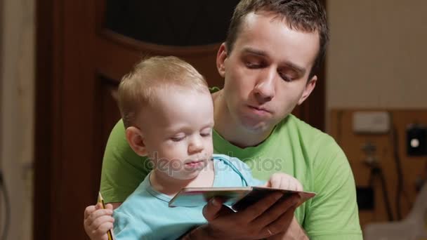 一个有吸引力的 2 岁男孩和父亲读一本书在家里。爸爸显示图片和儿子打电话来的动物. — 图库视频影像