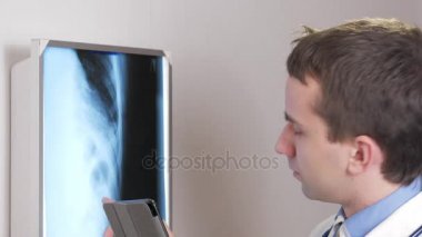 Genç bir doktor klinikte bir tablet bilgisayar kullanır. Bir erkek bir hasta röntgen sonuçları kaydeder.