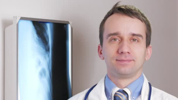 Молодой доктор смотрит в камеру и улыбается. На заднем плане висит рентген пациента. Рубашка с галстуком и стетоскопом на шее — стоковое видео
