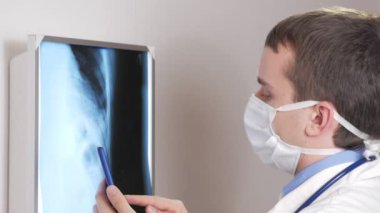 Bir yüz maskesi içinde genç bir doktor klinikte bir telefon kullanır. Bir erkek bir hasta röntgen sonuçları kaydeder.