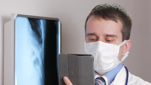 フェイス マスクで若い医師は、診療所でタブレット コンピューターを使用します。男が患者の x 線の結果を記録します。. — ストック動画