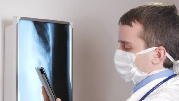 Un médico joven con una máscara facial usa una tableta en la clínica. Un hombre registra los resultados de una radiografía de pacientes . — Vídeos de Stock