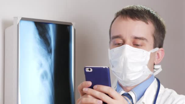 フェイス マスクで若い医師は、診療所で携帯電話を使用します。男が患者の x 線の結果を記録します。. — ストック動画