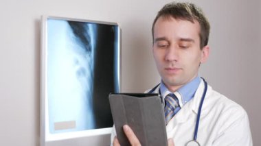 Genç bir doktor klinikte bir tablet bilgisayar kullanır. Bir erkek bir hasta röntgen sonuçları kaydeder.
