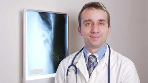 Ένας νεαρός γιατρός εξετάζει τη φωτογραφική μηχανή και χαμόγελα. Το φόντο που κρέμονται ακτινογραφία του ασθενούς. Πουκάμισο με μια γραβάτα και ένα στηθοσκόπιο στο λαιμό — Αρχείο Βίντεο