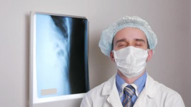 Bir yüz maskesi içinde genç bir doktor kamera ve gülümsüyor görünüyor. Koruyucu şapka kafasına. Hastanın x-ray asılı arka planı. Gömlek kravat ve bir steteskop boyun