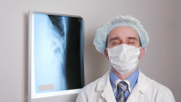 Ένας νεαρός γιατρός σε μια μάσκα προσώπου κοιτάζει την κάμερα και χαμόγελα. Προστατευτικό καπέλο στο κεφάλι. Το φόντο που κρέμονται ακτινογραφία του ασθενούς. Πουκάμισο με μια γραβάτα και ένα στηθοσκόπιο στο λαιμό — Αρχείο Βίντεο