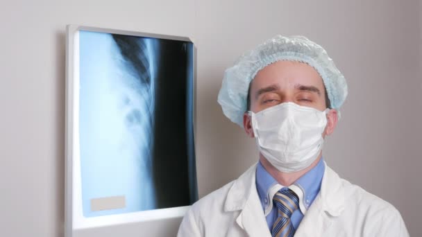 Een jonge dokter in een gezichtsmasker kijkt naar de camera en glimlach. Beschermende hoed op het hoofd. Tegen de achtergrond opknoping x-ray van de patiënt. Shirt met een gelijkspel en een stethoscoop op de hals — Stockvideo