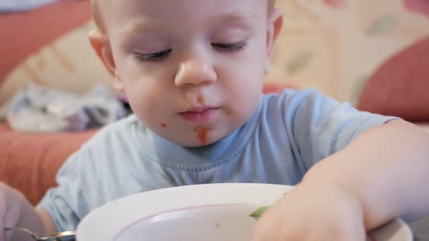 Atrakcyjny chłopiec 2 lat jest jedzenie zupy czerwony, sam. Liść laurowy złowionych w płycie i dziecko bawi się z nim. — Wideo stockowe