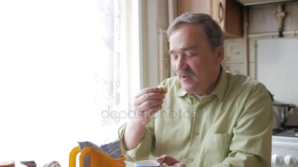 Ένας ηλικιωμένος άντρας με μουστάκι κατέχει ένα φλιτζάνι ζεστό ρόφημα και ποτά. Κάθεται κοντά στο παράθυρο και έχει το πρωινό στο σπίτι — Αρχείο Βίντεο