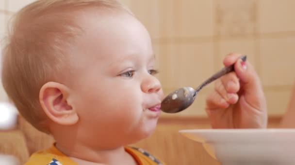 Baby 2 jaar oud eet PAP met een lepel. Moeder zit naast de jongen. woninginrichting — Stockvideo