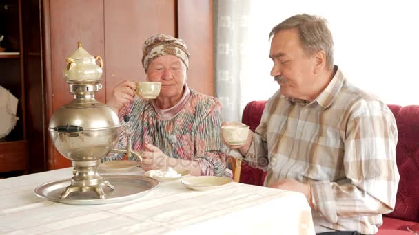 An elderly couple is drinking tea from a vintage Russian kettle samovar. A man with a mustache talking with his wife in a kerchief — Stock Video