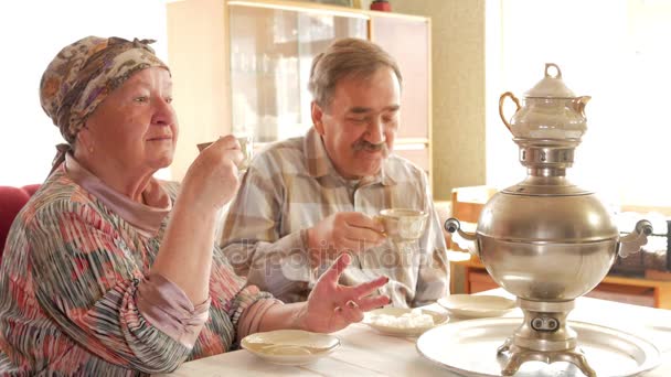 Um casal de idosos está bebendo chá de um samovar de chaleira russo vintage. Um homem com bigode a falar com a mulher num lenço — Vídeo de Stock