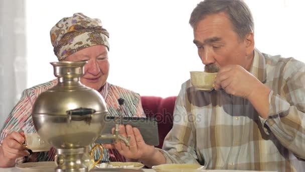 Elderly couple drink tea from a vintage Russian teapot samovar and watch a photo in the tablet. A man with a mustache and a woman discussing the image. — Stock Video