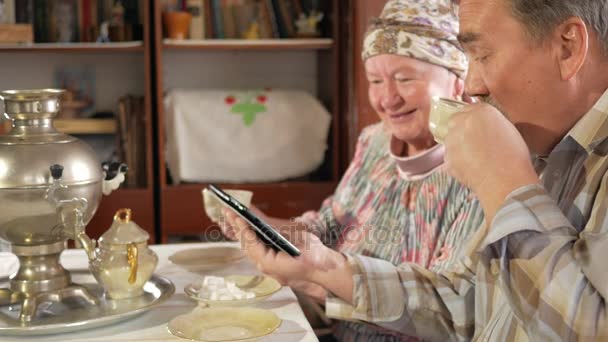 Ein älteres Ehepaar trinkt Tee aus einer alten russischen Teekanne Samowar und schaut sich ein Foto in der Tablette an. ein Mann mit Schnurrbart und eine Frau diskutieren das Bild. — Stockvideo
