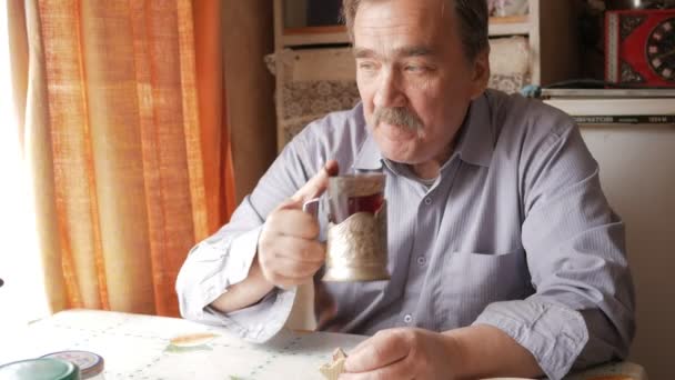 Пожилой человек с усами держит чашку горячего напитка и напитков. Он сидит у окна и завтракает дома. — стоковое видео