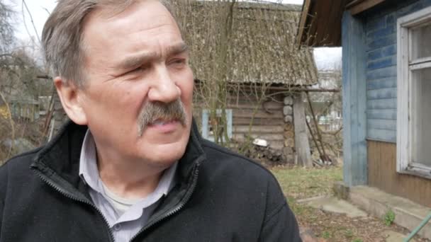 An elderly man with a mustache smokes in the street near the old house. Dressed in a black sweater — Stock Video